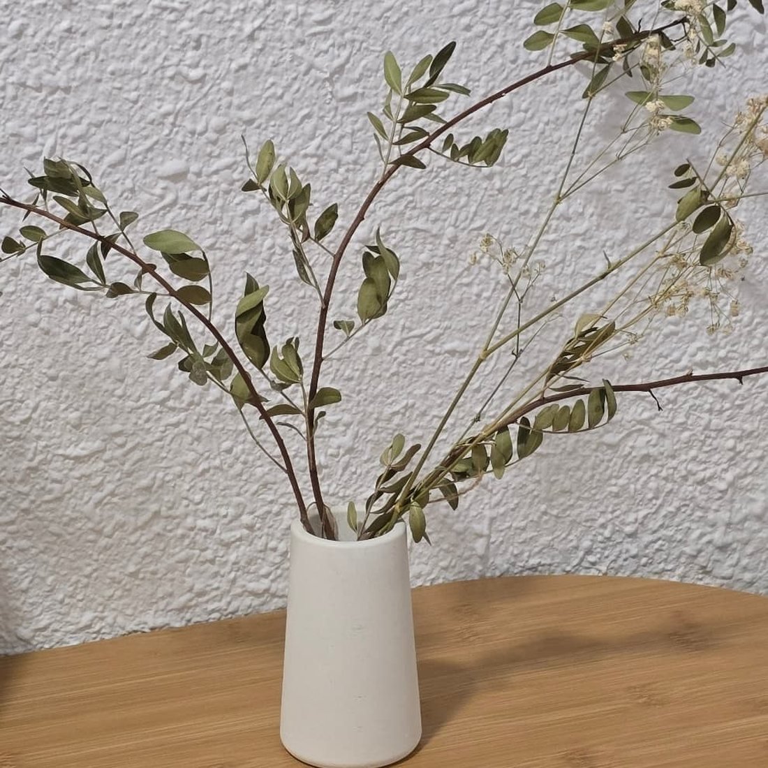 Florero cónico de cemento de Made Taxa con ramas secas sobre una mesa de madera